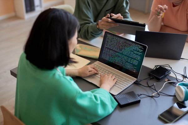 Young female student typing on laptop keyboard while decoding data on screen against two classmates or colleagues discussing presentation